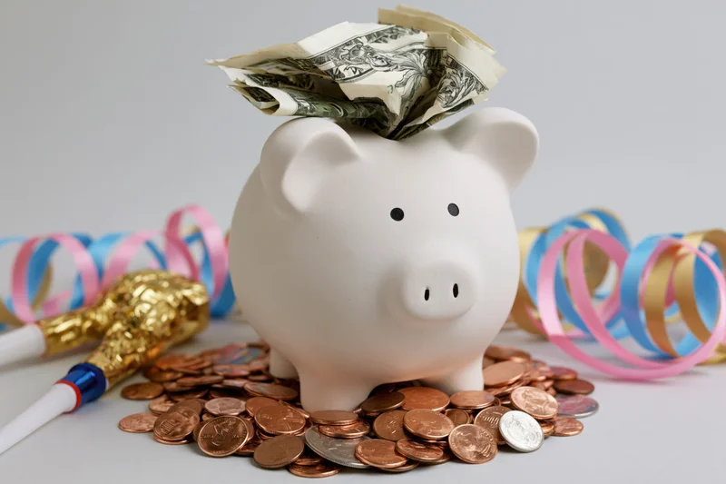 White ceramic piggy bank stuffed with folded us dollar bills surrounded by coins and party streamers on a plain surface