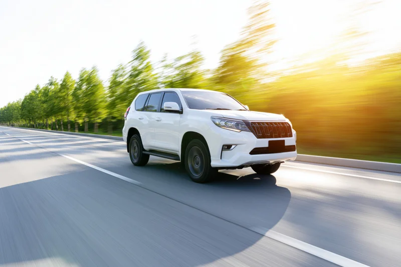 White midsize suv driving at speed on a two-lane road with blurred trees and sunlight in the background