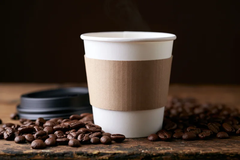 White paper takeaway coffee cup with cardboard sleeve and steam rising, surrounded by roasted coffee beans on wooden surface