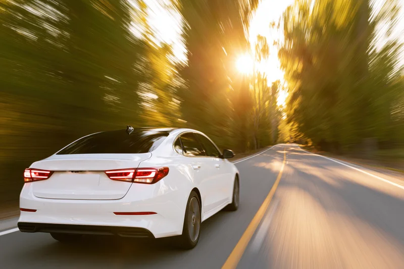 White sedan driving away on a two-lane country road lined with trees at golden hour