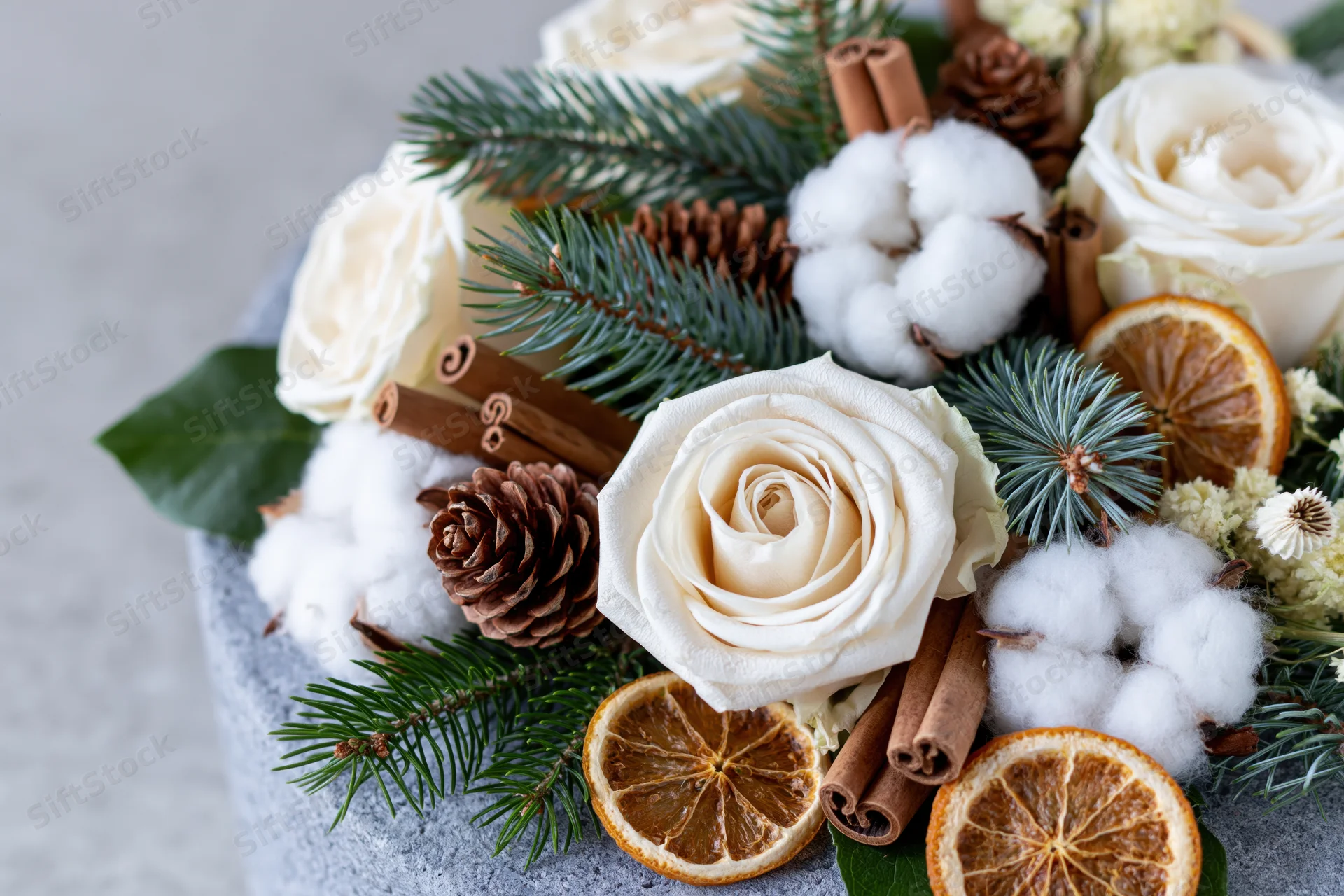 Winter floral arrangement with white roses, pine branches, pinecones, cinnamon sticks, dried orange slices, and cotton fluff on a gray stone surface