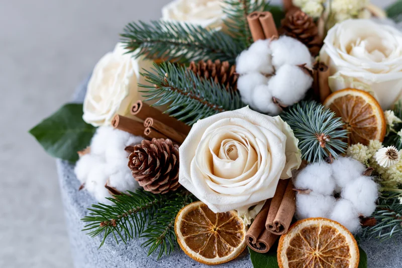 Winter floral arrangement with white roses, pine branches, pinecones, cinnamon sticks, dried orange slices, and cotton fluff on a gray stone surface