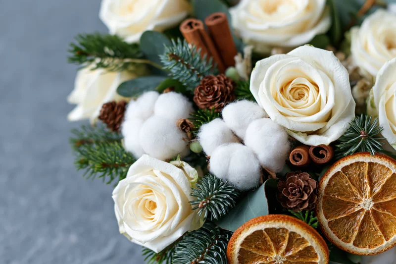 Winter floral arrangement with white roses, cotton, pine, cinnamon, and dried oranges on grey stone surface