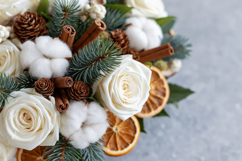 Winter floral arrangement with white roses, cotton, pine, cinnamon sticks and dried oranges on gray stone surface