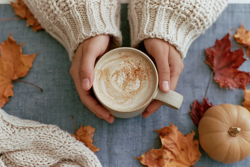 Woman’s hands in a cream knit sweater cradling a ceramic mug of latte with cinnamon on a blue fabric surface surrounded by autumn leaves and a small pumpkin
