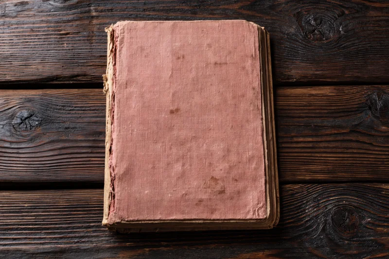 Worn antique book with blank faded pink cover lying flat on dark wooden surface
