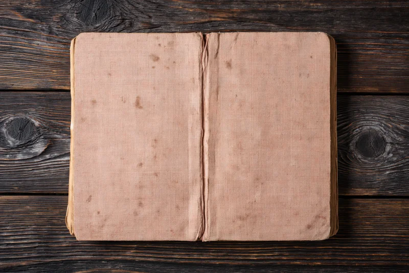 Worn antique book lying flat open on dark wooden surface with blank faded cover facing up