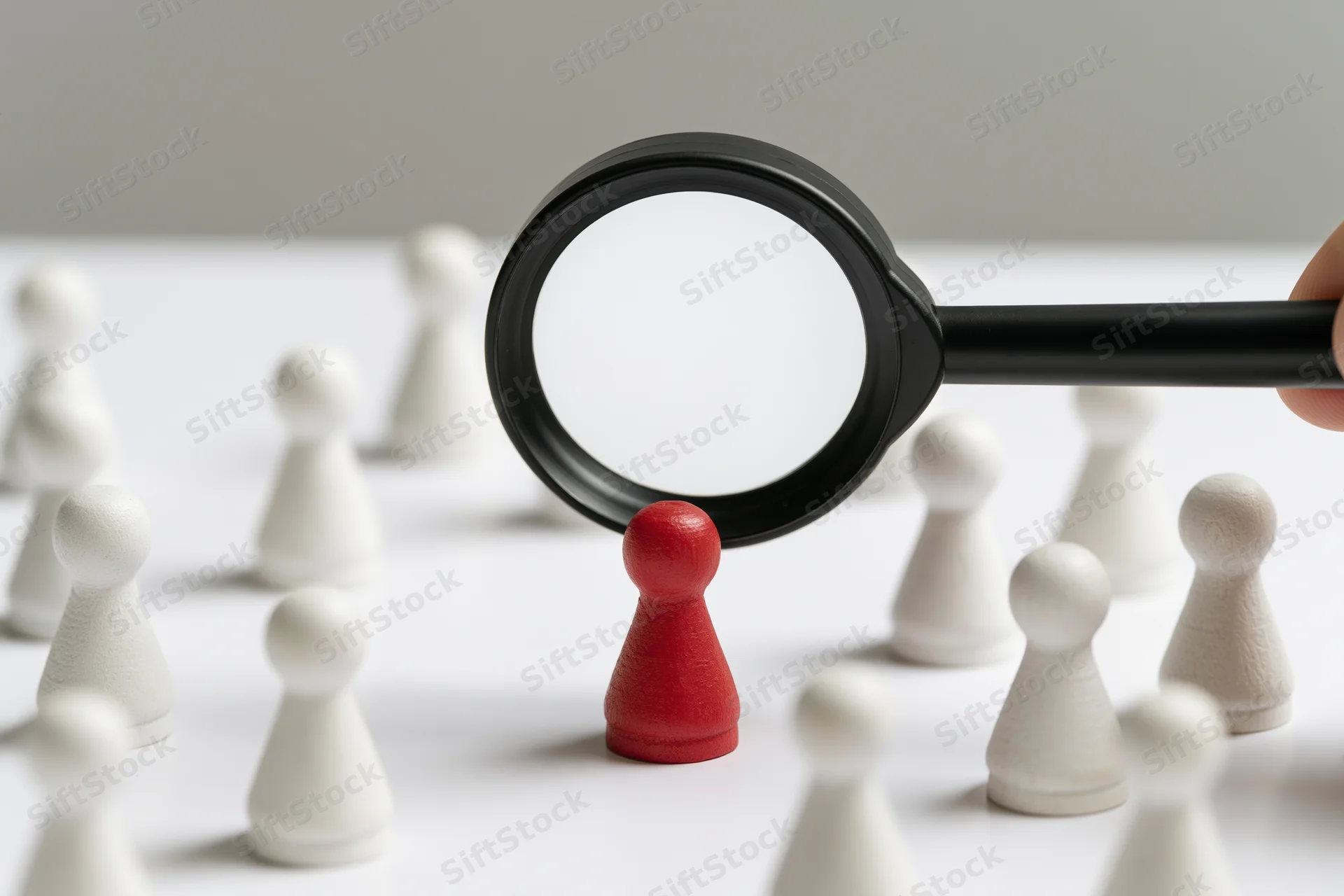 Hand holding magnifying glass focusing on red figurine among white ones on white surface