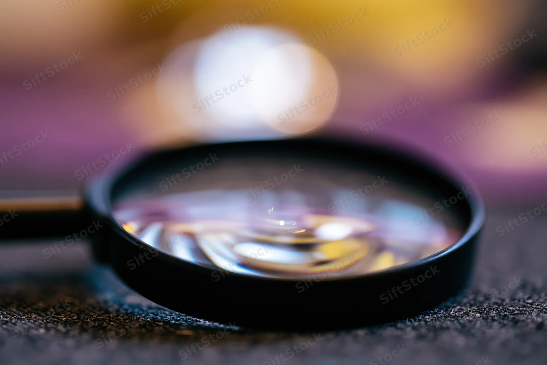 Black magnifying glass resting on dark textured surface with blurred background and light reflections in lens