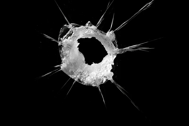 Bullet hole in a glass pane with radiating cracks spreading outward against black background