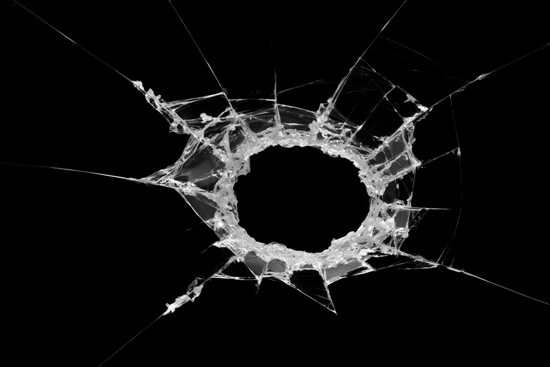 Bullet hole in shattered glass pane centered in frame with radial cracks against black background