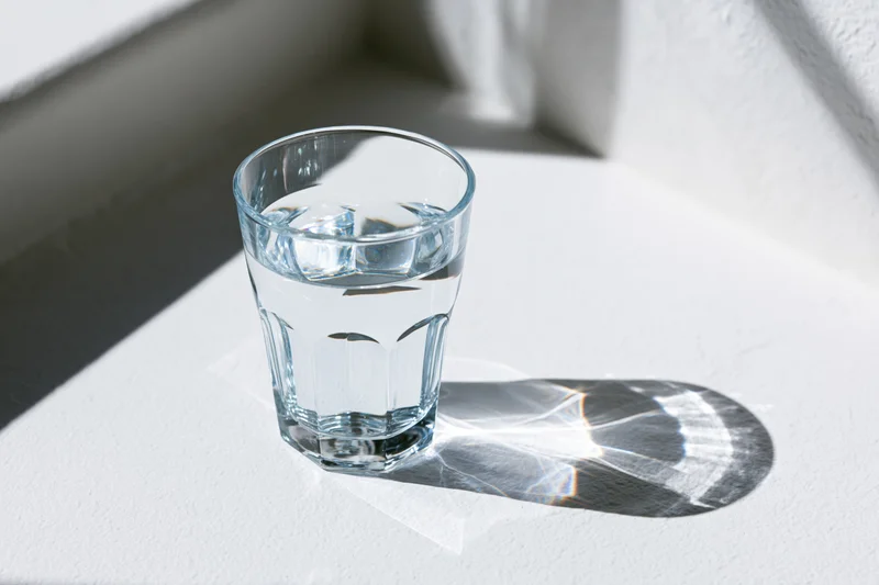 Faceted glass filled with water sitting on white surface in sunlight casting sharp shadow and light refraction patterns