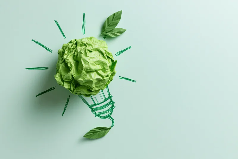 Crumpled green paper ball shaped as a lightbulb with drawn filament and leaves on a plain background