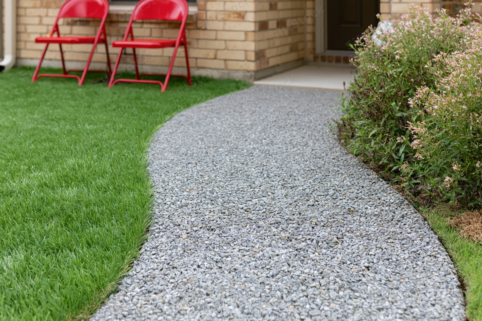 Curved gravel pathway winding through a front yard with green lawn and red folding chairs near a brick house entrance