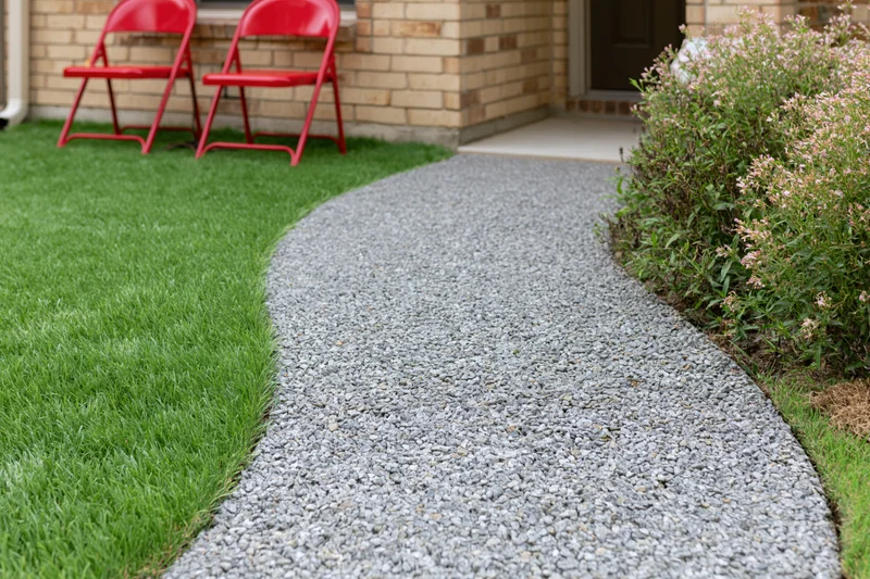 Curved gravel pathway winding through a front yard with green lawn and red folding chairs near a brick house entrance