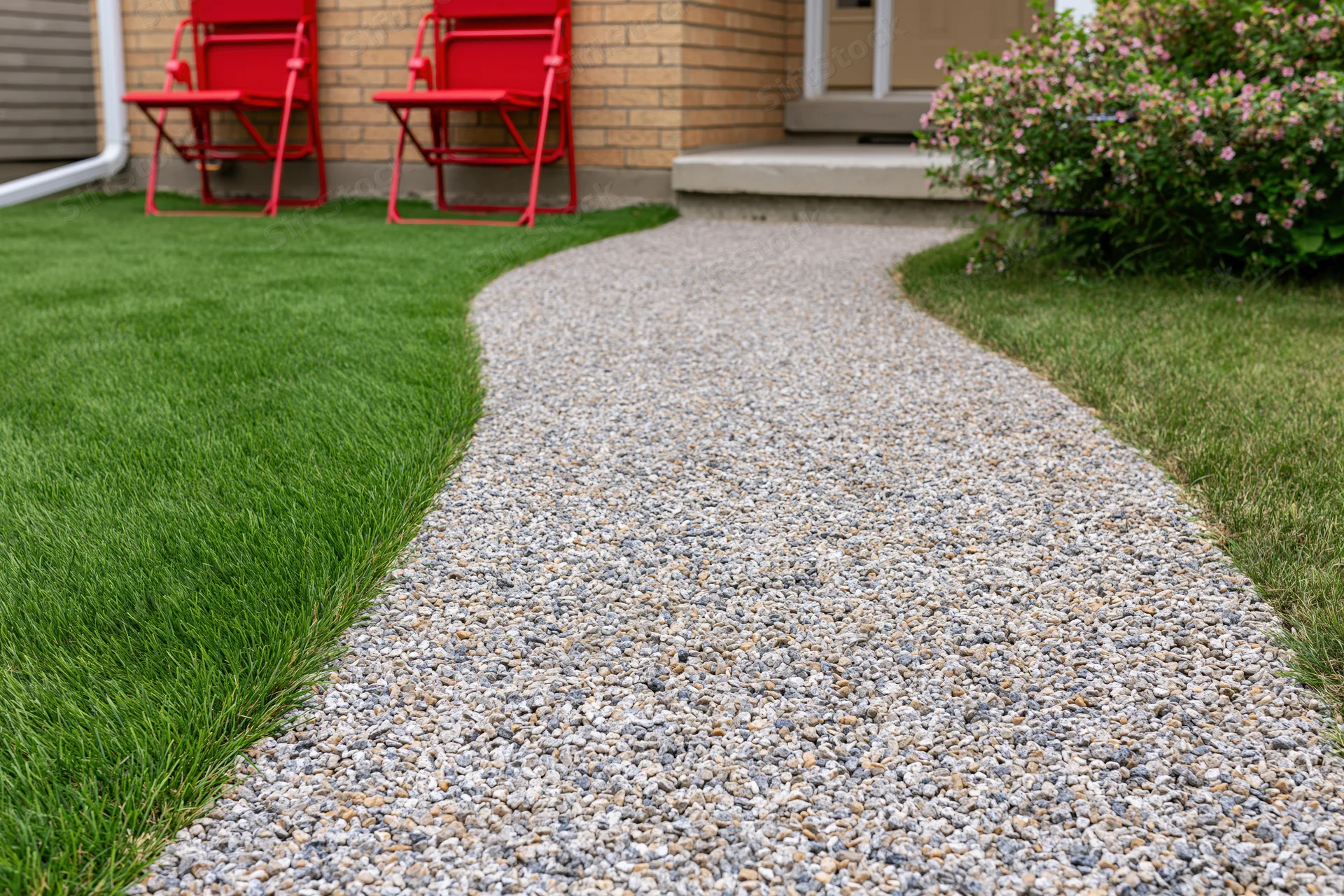 Curved gravel pathway winding through a front yard with green lawn and red folding chairs near a brick house entrance