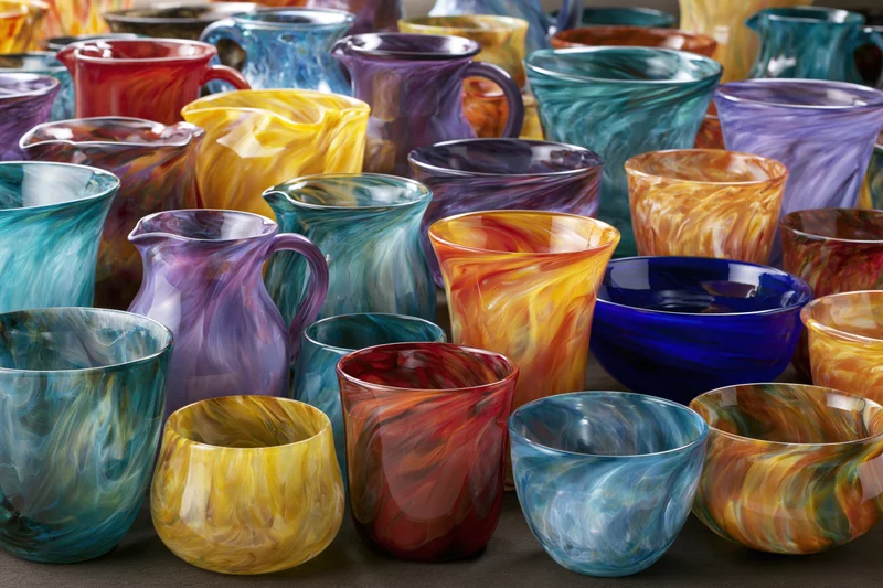 Hand-blown glass vases pitchers and bowls arranged in a dense display on a dark surface