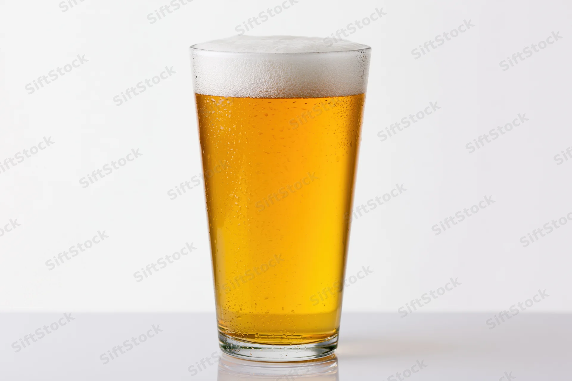 Full pint glass of pale lager with white foam head on white surface