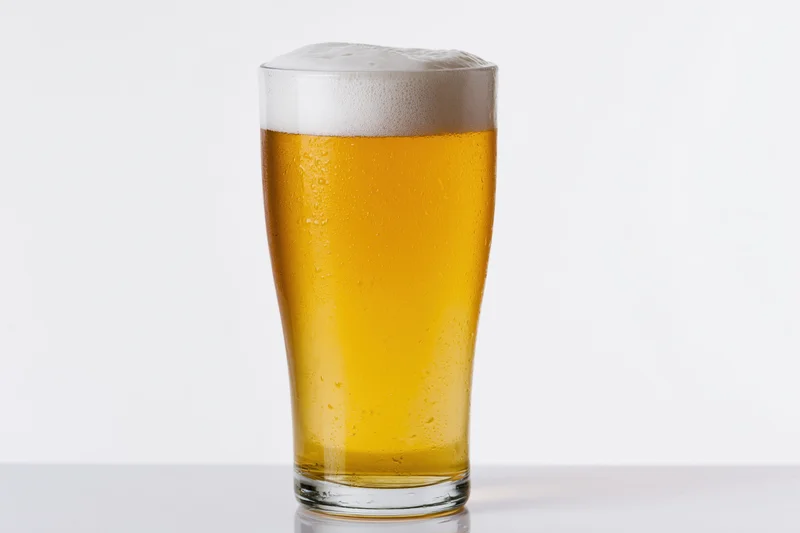 Full pint glass of pale lager with white foam head on white surface