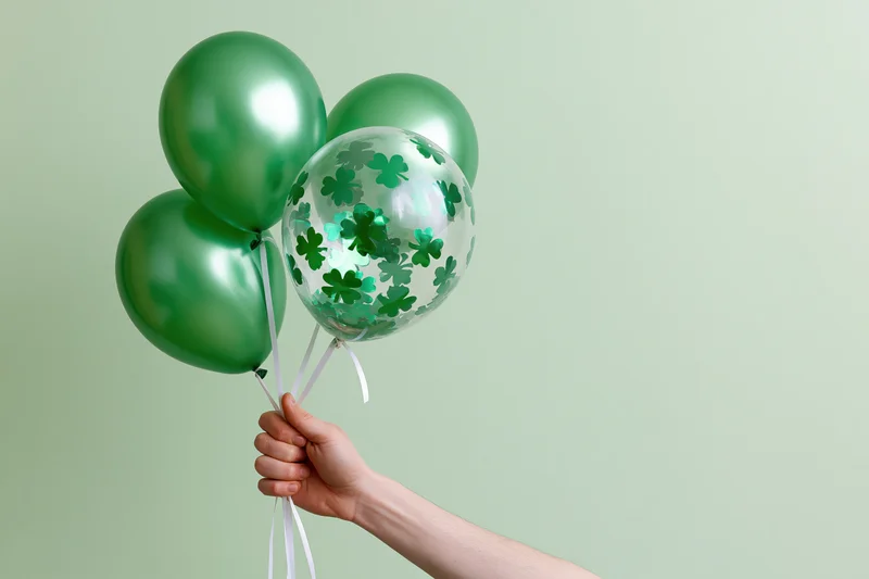 Hand holding green balloons with shamrock confetti against light green background