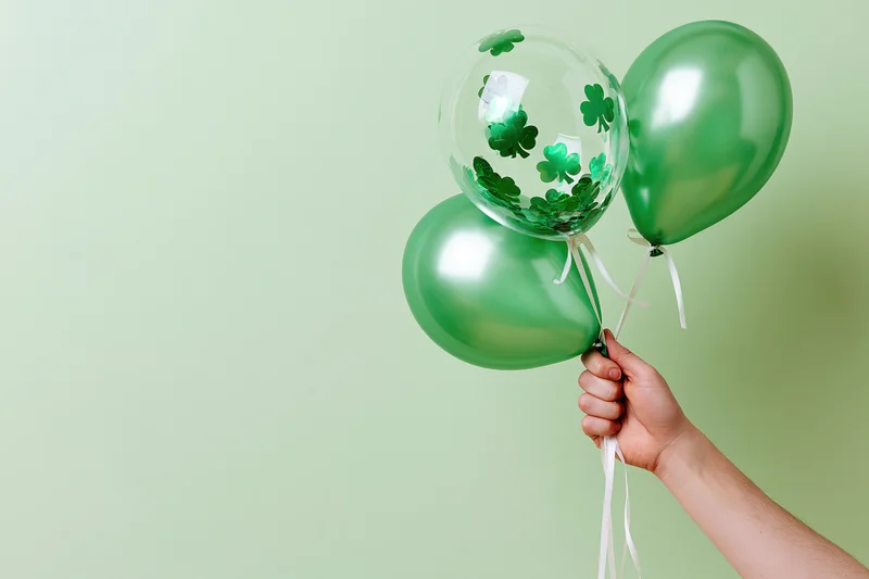 Hand holding three green balloons with shamrocks against a plain light green background
