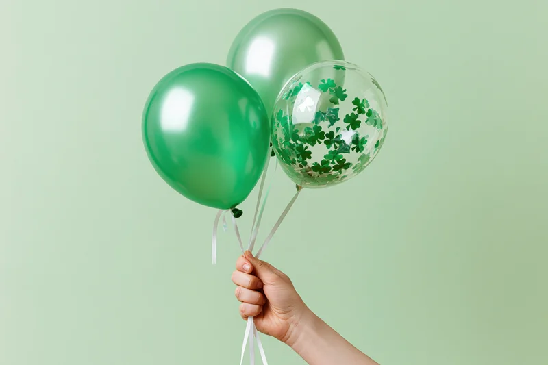 Hand holding three green balloons with shamrock confetti against light green background