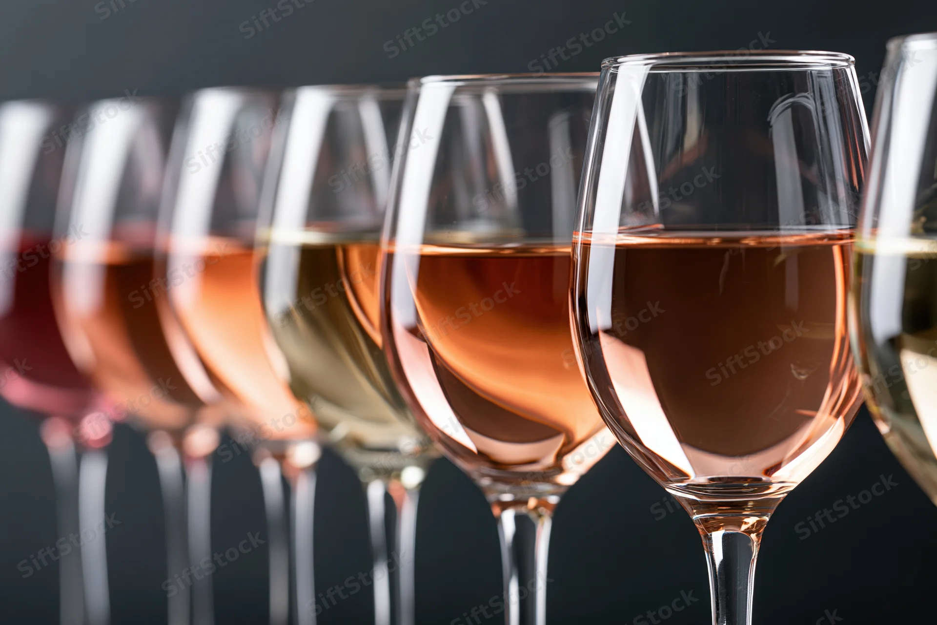 Row of stemmed wine glasses filled with varying shades of wine arranged in a line against dark background