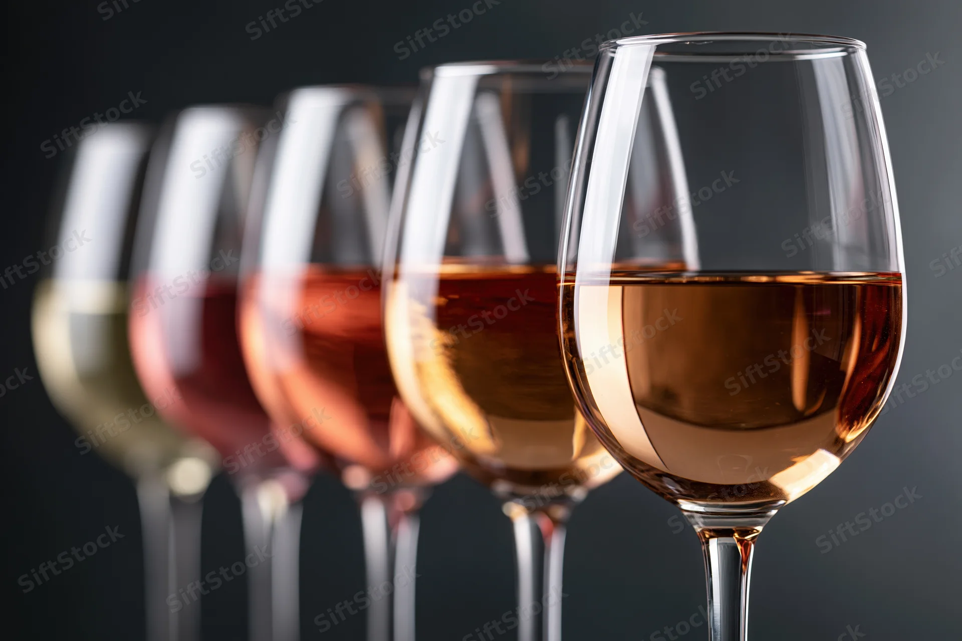 Row of stemmed wine glasses filled with varying shades of wine arranged in a line against dark background