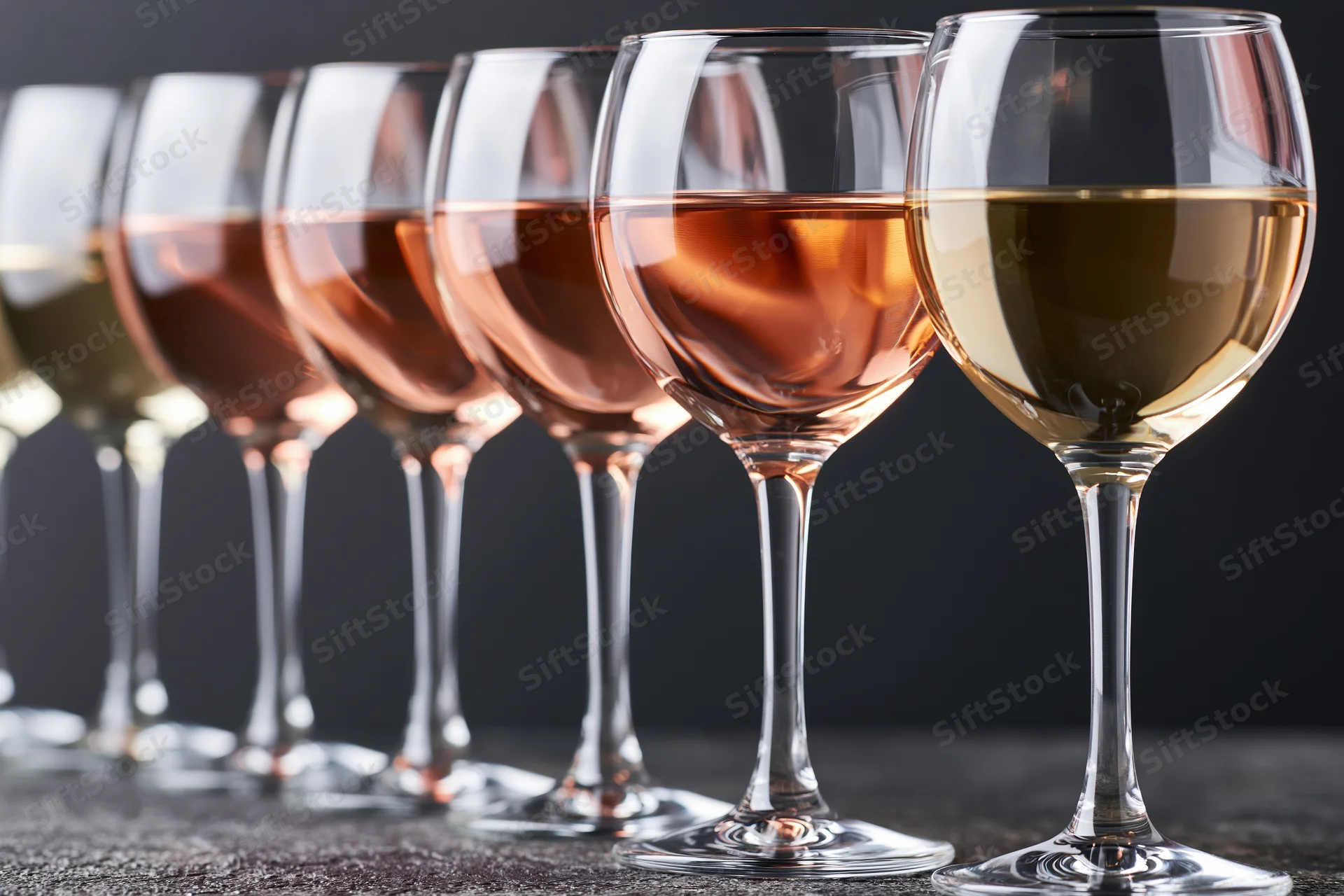 Row of stemmed wine glasses filled with varying shades of wine arranged on dark surface