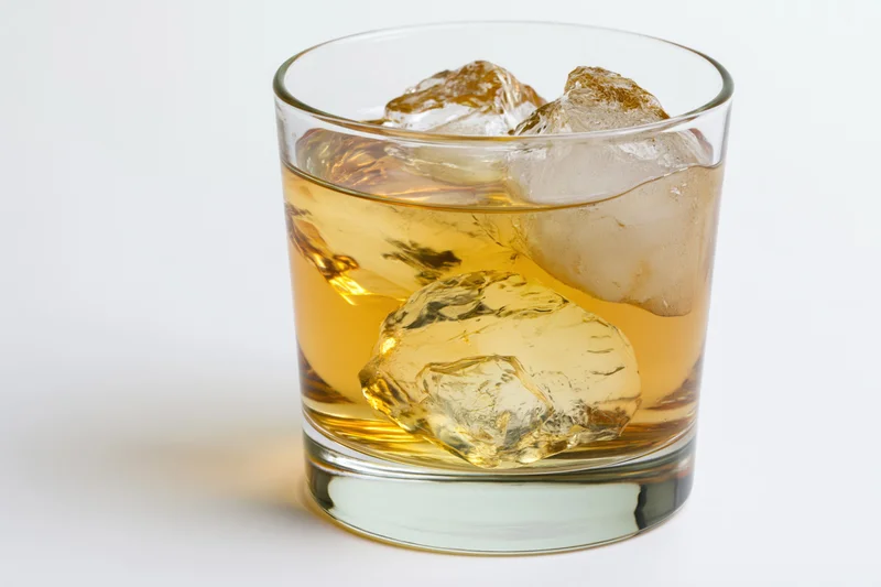 Short rocks glass filled with amber whiskey and large ice cubes on white background