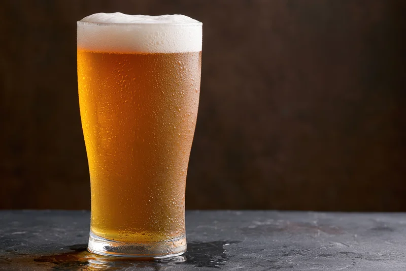 Tall pint glass filled with cold lager beer covered in condensation on dark surface