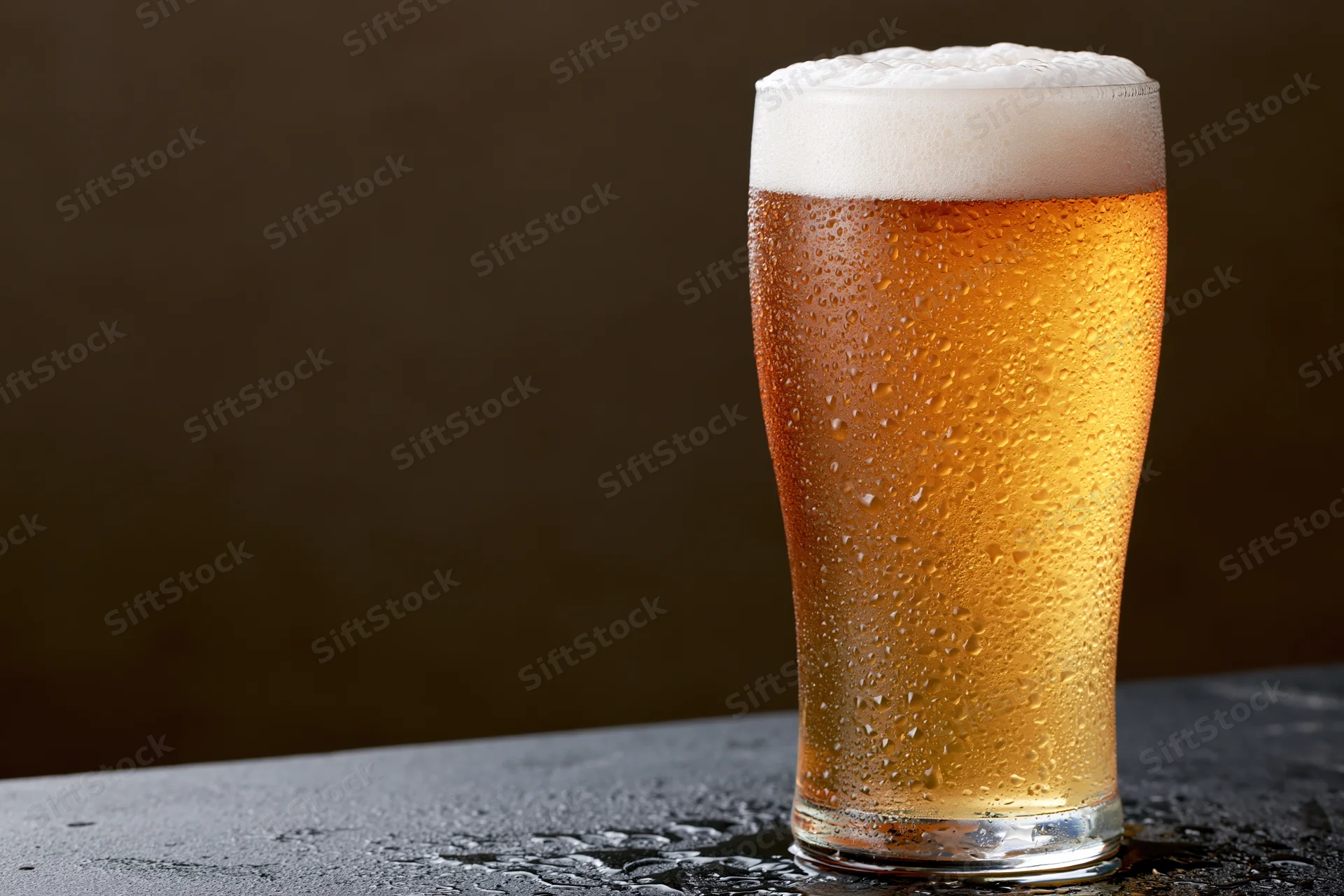 Tall pint glass filled with cold lager beer covered in condensation on dark surface