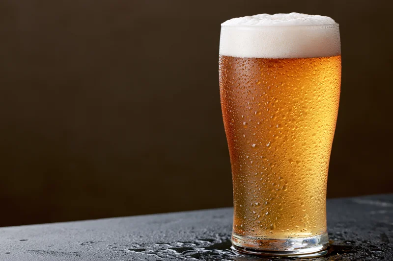 Tall pint glass filled with cold lager beer covered in condensation on dark surface