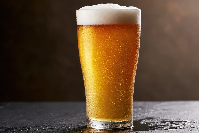 Tall pint glass filled with cold lager beer covered in condensation on dark surface