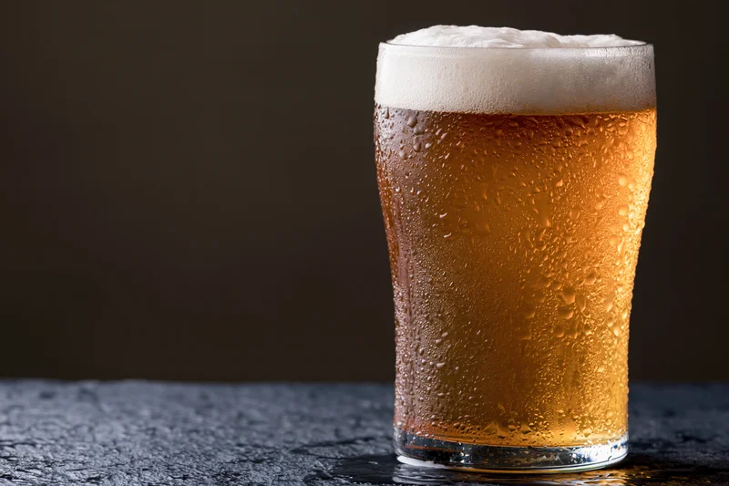 Tall pint glass filled with cold lager beer covered in condensation on dark textured surface