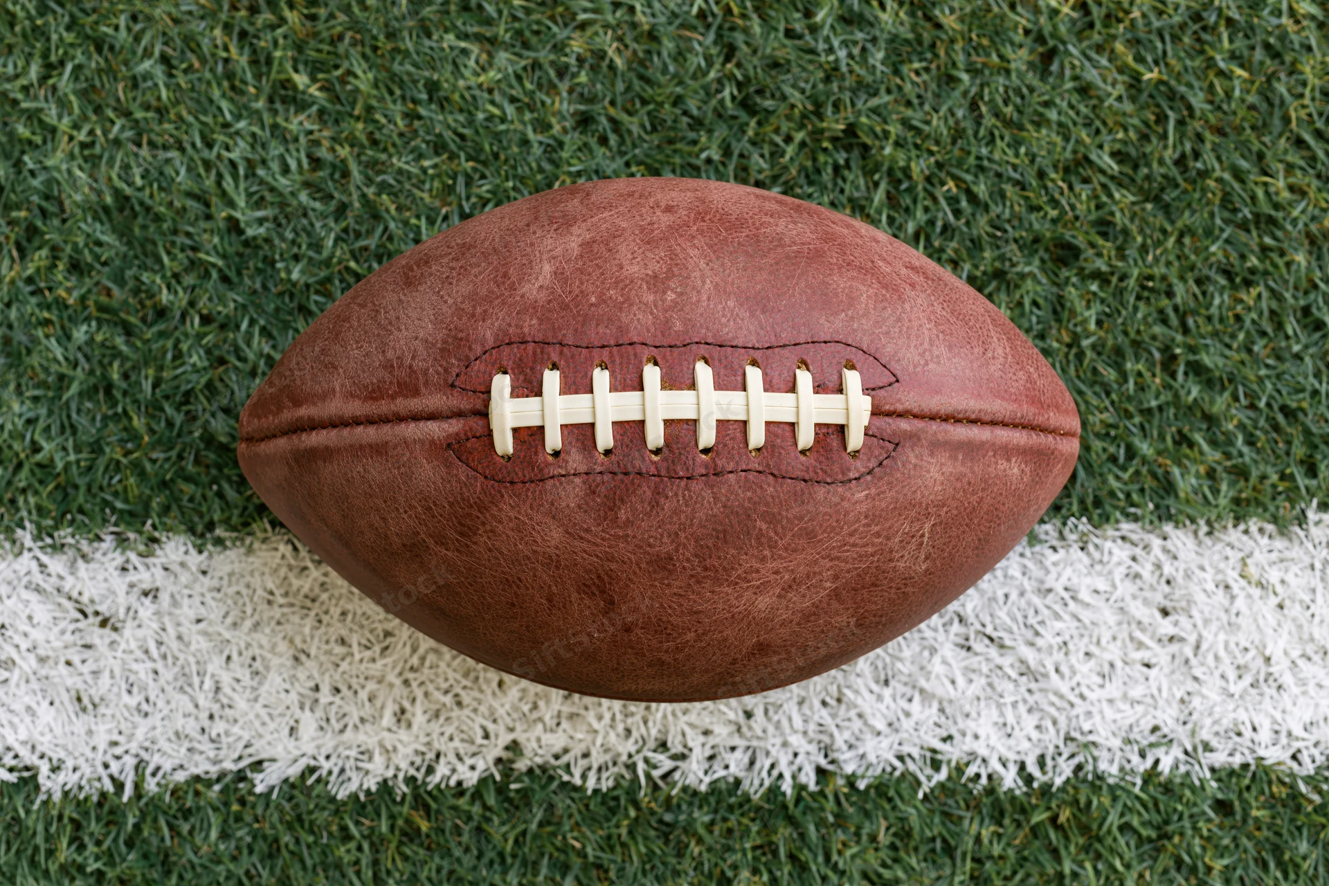 American football resting on a painted white yard line on green turf field