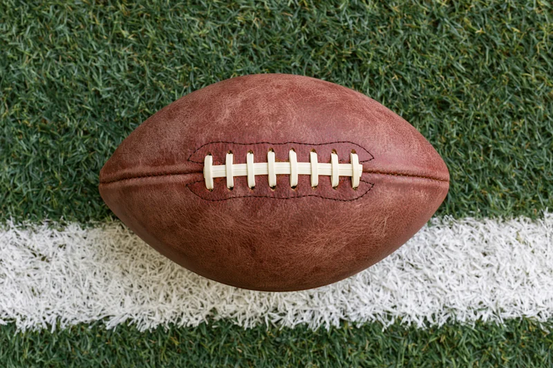 American football resting on a painted white yard line on green turf field