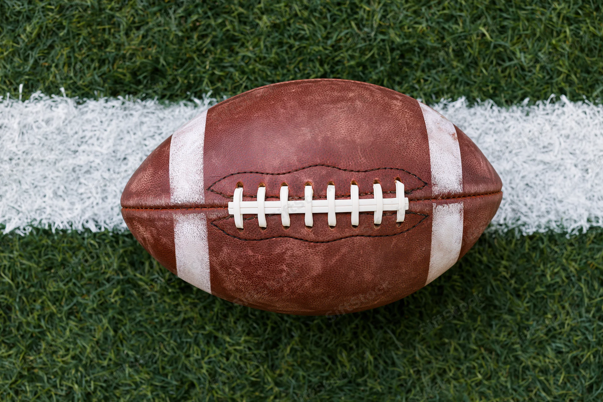 American football resting on painted white yard line on green turf field