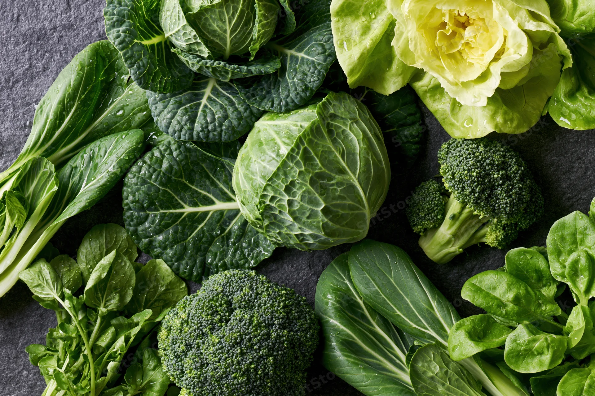 Assorted green vegetables including broccoli, savoy cabbage, and bok choy arranged on a dark stone surface