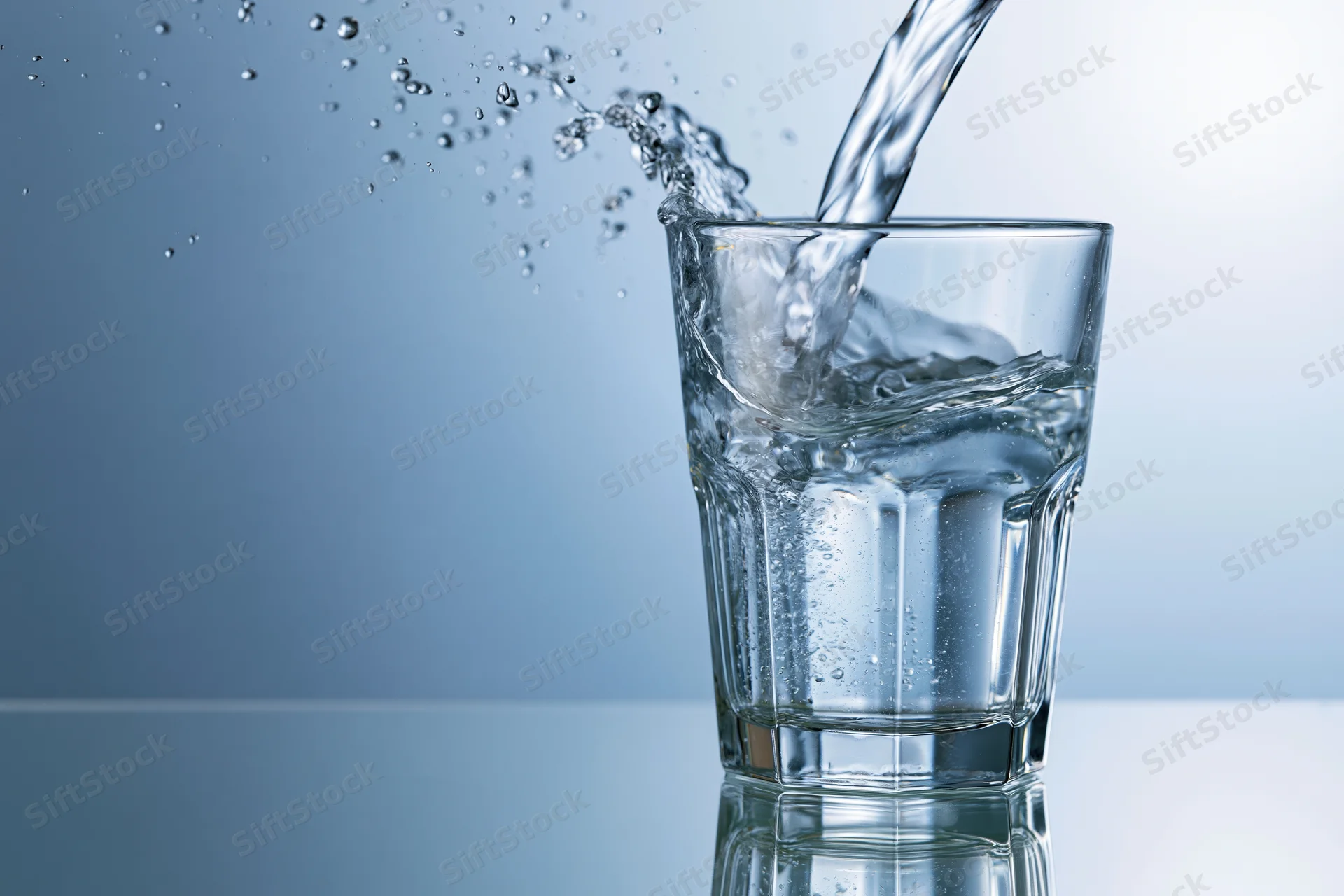 Clear glass tumbler being filled with water mid-pour splash on reflective surface