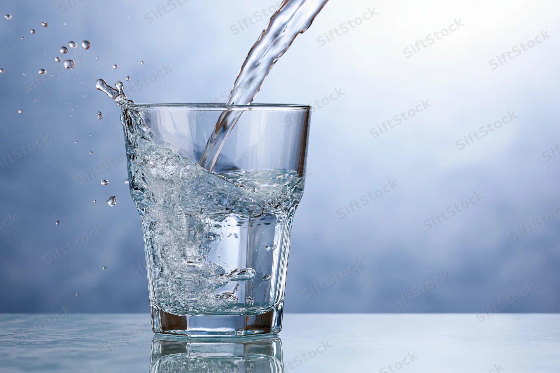 Clear glass tumbler being filled with water mid-pour splash on reflective surface