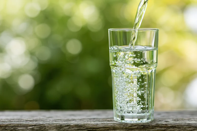 Clear water being poured into a tall glass on a wooden table outdoors