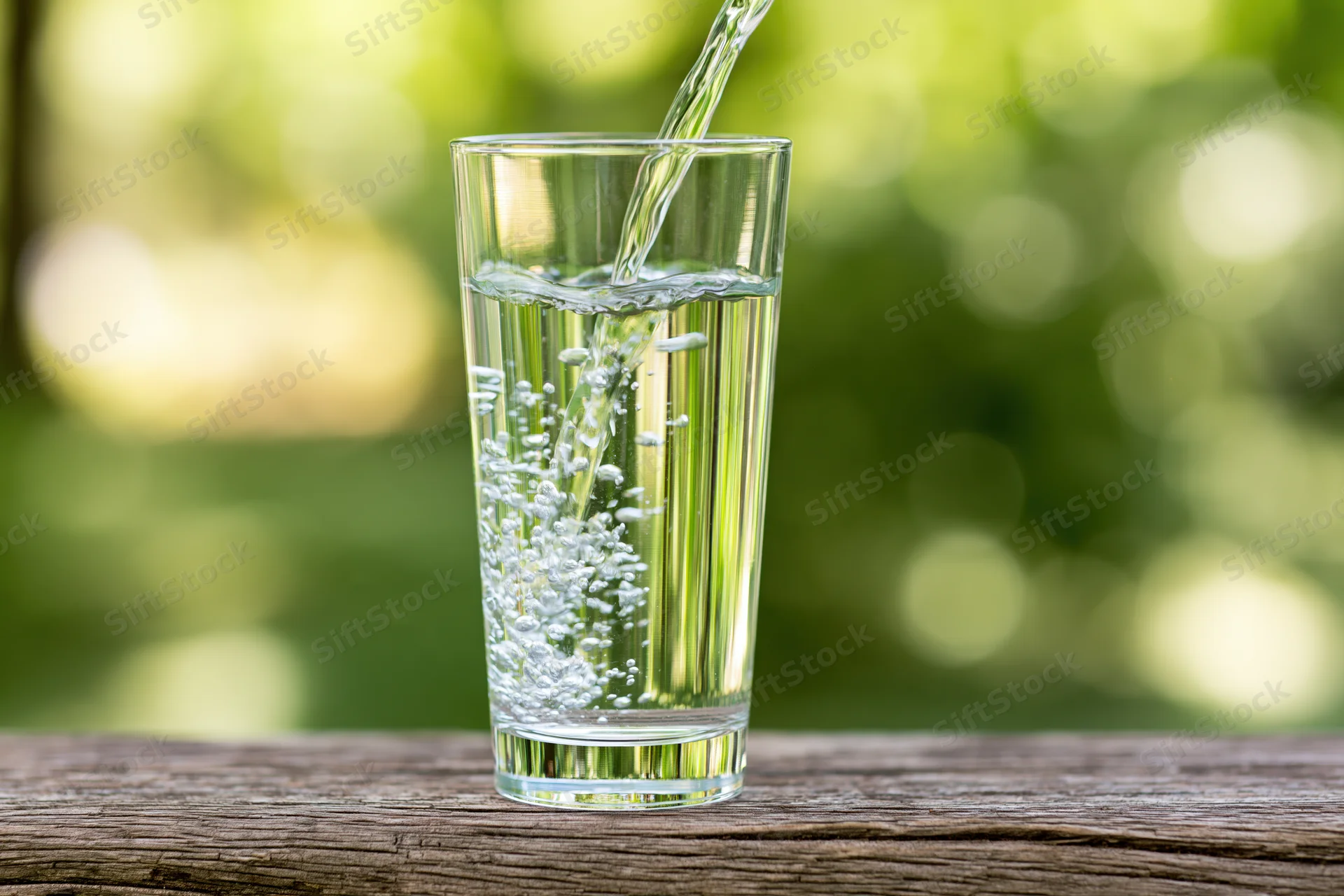 Clear water being poured into a tall glass on a wooden table outdoors with green foliage in the background
