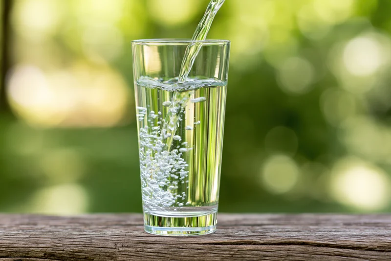 Clear water being poured into a tall glass on a wooden table outdoors with green foliage in the background