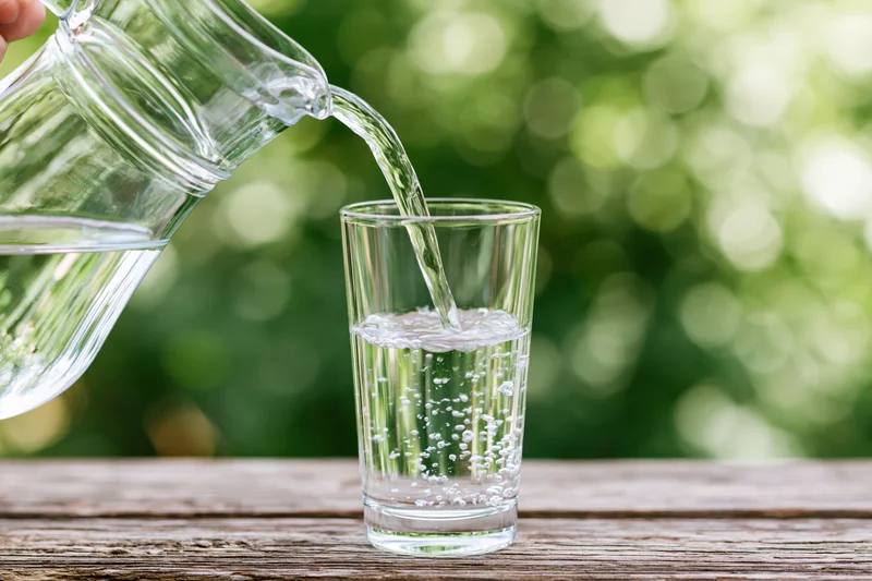 Clear water pouring from a glass pitcher into a tall drinking glass on a wooden table outdoors