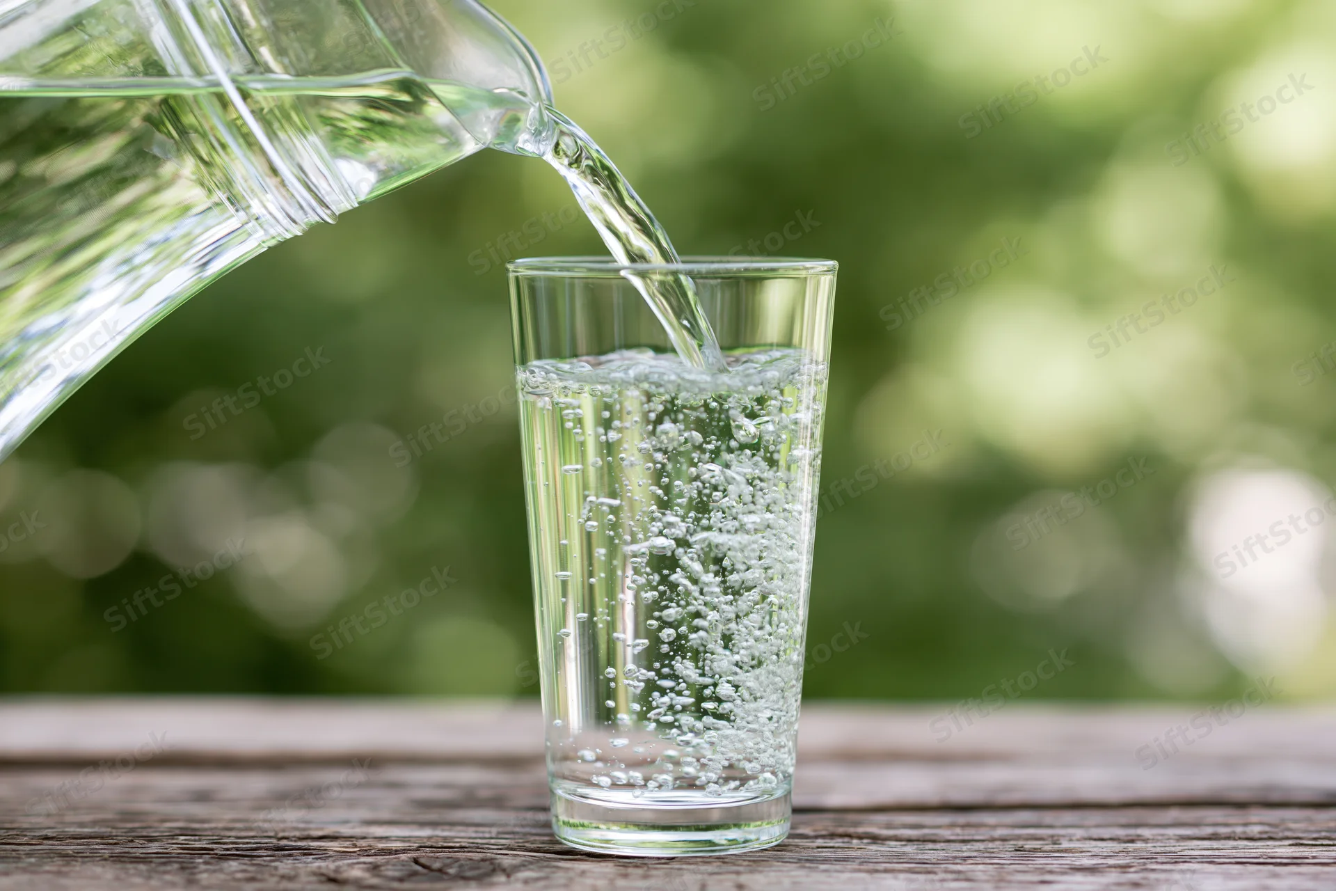 Clear water pouring from a glass pitcher into a tall drinking glass on a wooden table outdoors