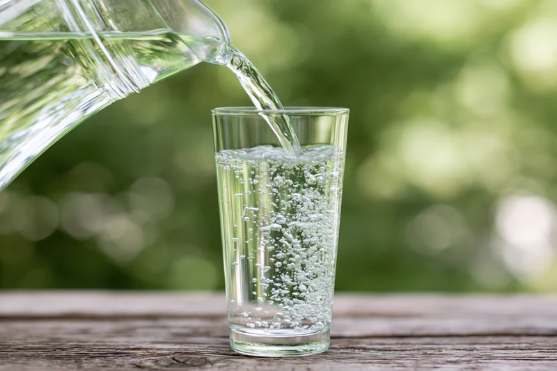 Clear water pouring from a glass pitcher into a tall drinking glass on a wooden table outdoors