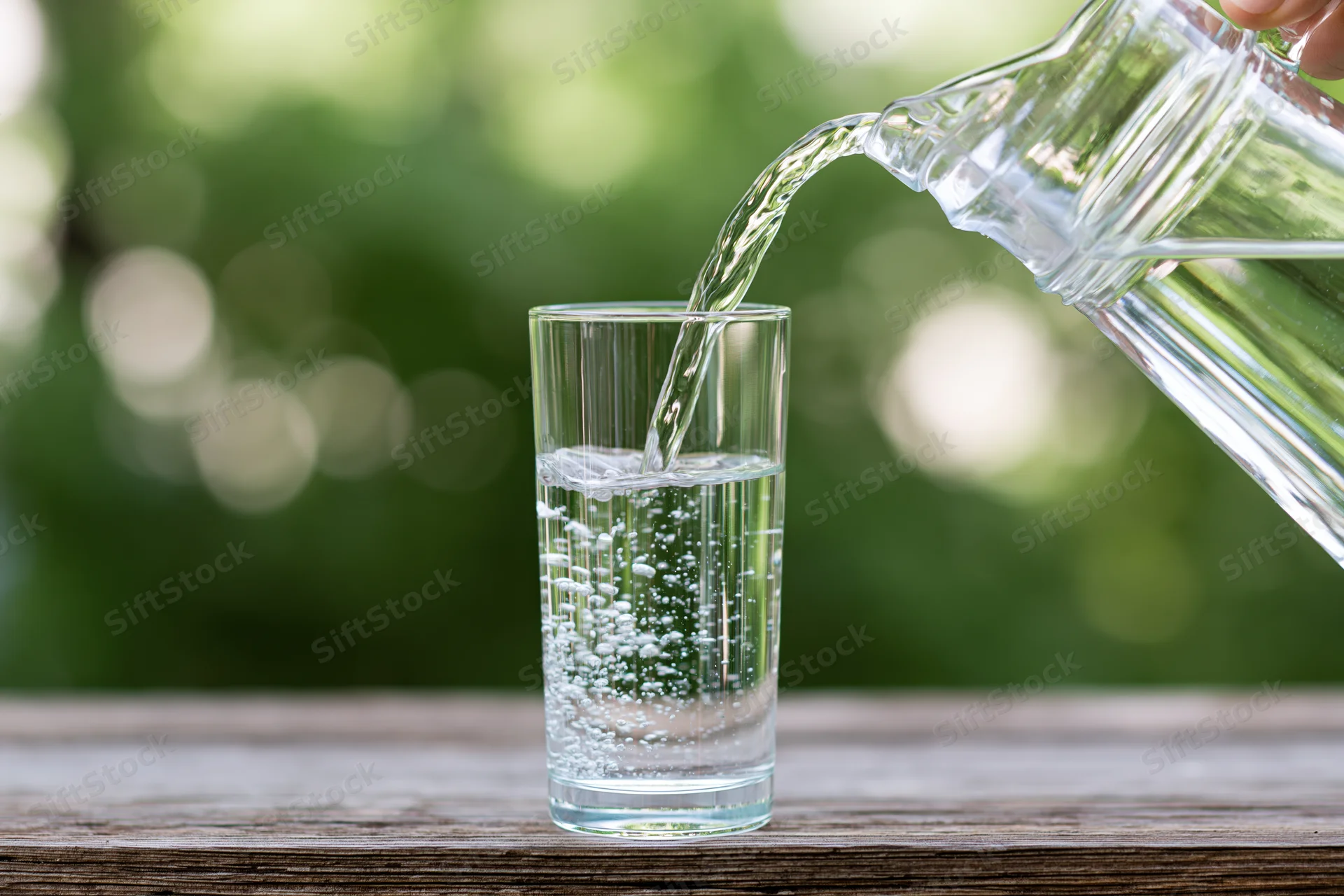 Clear water pouring from a glass pitcher into a tall drinking glass on a wooden table outdoors