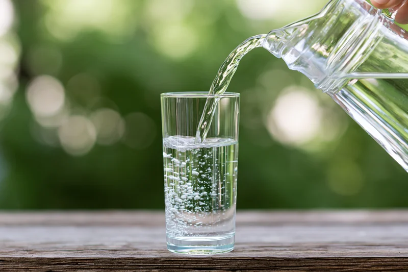 Clear water pouring from a glass pitcher into a tall drinking glass on a wooden table outdoors