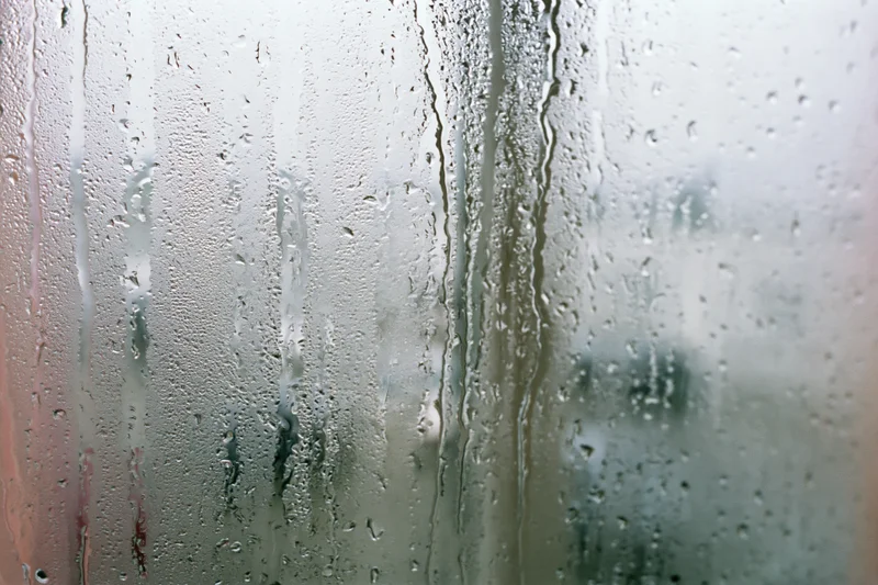 Fogged window glass covered in rain droplets and condensation streaks, showing blurred outdoor view through wet surface
